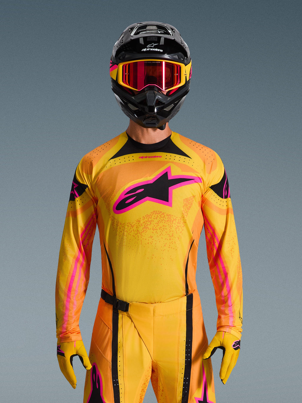 Alpinestars Maglia Techstar Nomur 2026 in arancione, giallo e rosa, indossata da un pilota di motocross con casco nero e occhiali rosa abbinati, con un grande logo Alpinestars rosa sul petto, vestibilità atletica da gara per prestazioni off-road