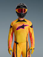 Alpinestars Maglia Techstar Nomur 2026 in arancione, giallo e rosa, indossata da un pilota di motocross con casco nero e occhiali rosa abbinati, con un grande logo Alpinestars rosa sul petto, vestibilità atletica da gara per prestazioni off-road