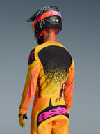 Maglia Alpinestars Techstar Nomur 2026 in Arancione Giallo Rosa, vista posteriore di un pilota di motocross con attrezzatura completa, pantaloni coordinati e casco nero, design vivace con accenti gialli e rosa e pannello posteriore nero, vestibilità atletica da gara per prestazioni off-road
