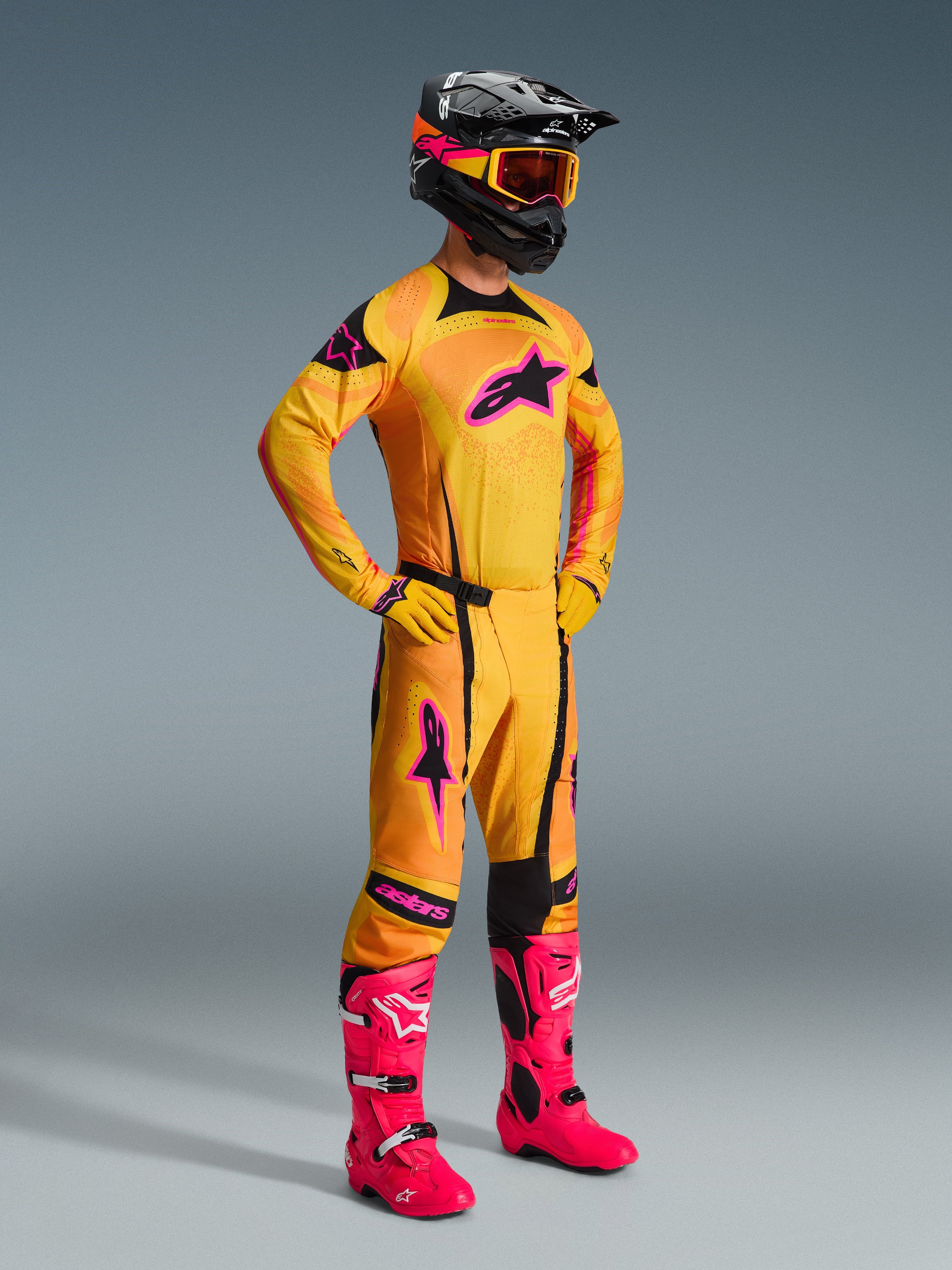Pilota di motocross che indossa la maglia Techstar Nomur 2026 in arancione, giallo e rosa, pantaloni arancioni coordinati con dettagli rosa, stivali rosa acceso, guanti gialli e casco nero con occhiali gialli, set completo di abbigliamento da motocross coordinato