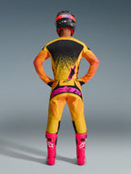 Pilota di motocross visto di spalle che indossa la maglia Alpinestars Techstar Nomur 2026 in Arancione Giallo Rosa, abbigliamento da gara coordinato con pantaloni abbinati con logo rosa e stivali rosa acceso, vestibilità atletica da gara per prestazioni fuoristrada di livello avanzato.