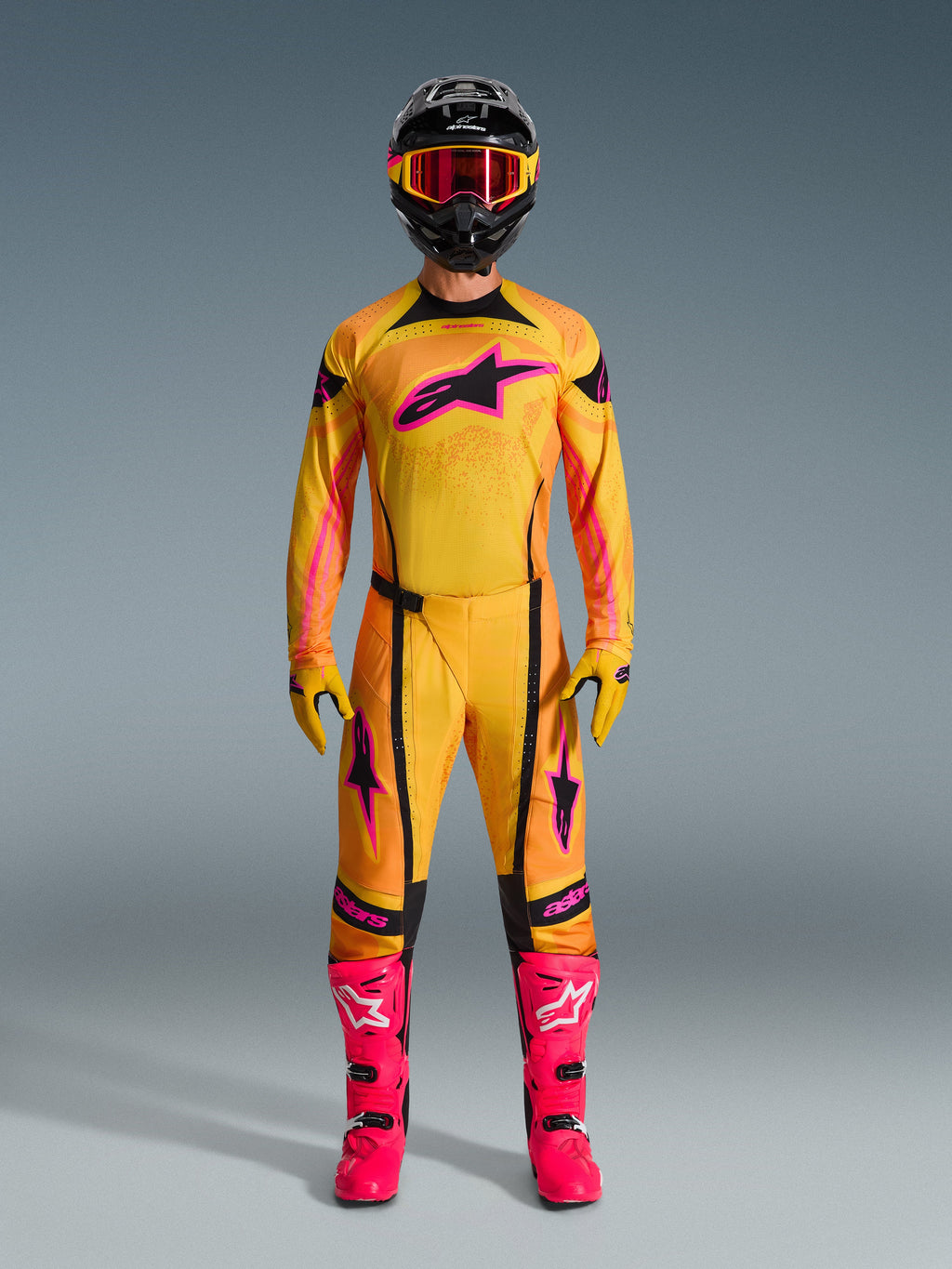 Set completo motocross 2026 Techstar Nomur, abbigliamento da gara completo indossato da un pilota, maglia con sfumatura giallo e arancione e logo stella rosa, pantaloni e guanti coordinati, stivali rosa acceso e casco nero, kit MX professionale su sfondo grigio