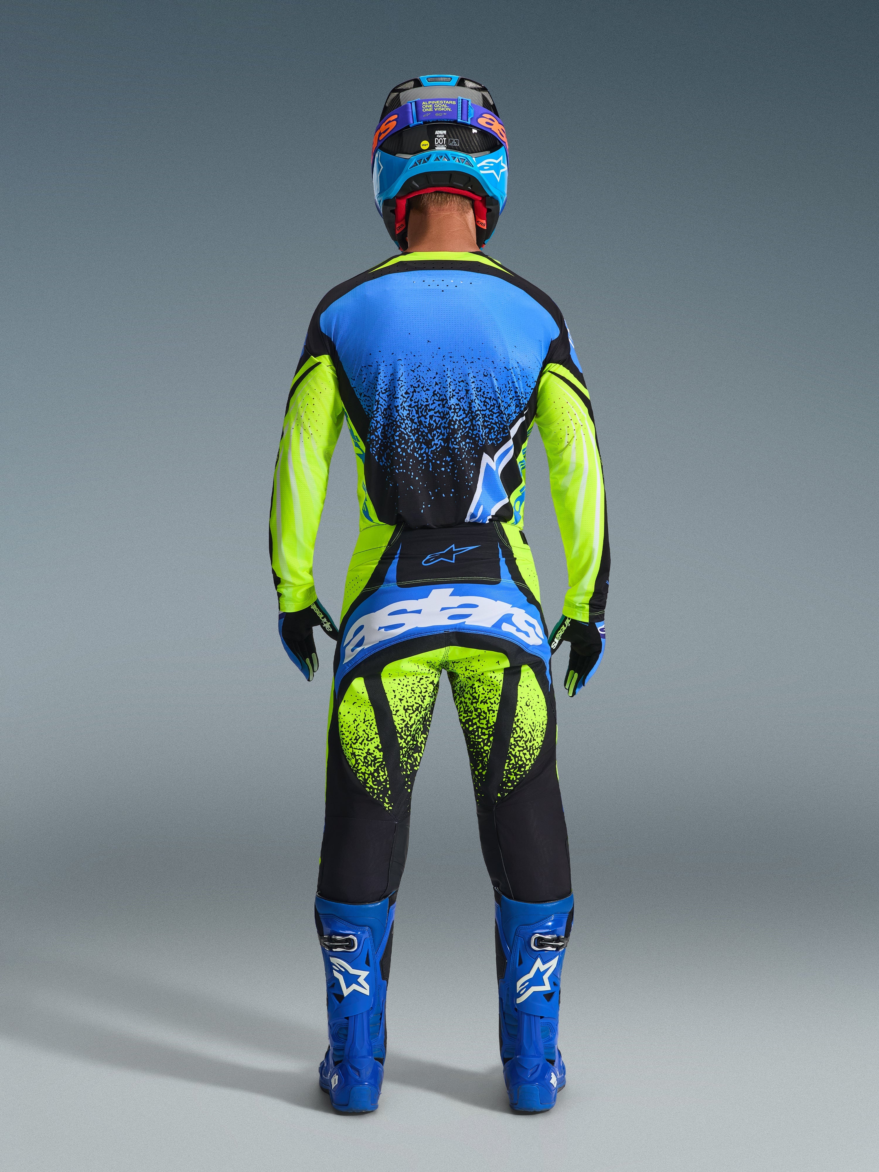 Vista posteriore di un pilota con il completo Alpinestars 2026 Techstar Nomur, maglia blu e giallo neon con accenti neri a effetto spray, pantaloni coordinati con grande logo 'astars' blu sulla seduta, stivali blu con loghi bianchi e casco blu e nero, abbigliamento professionale da motocross