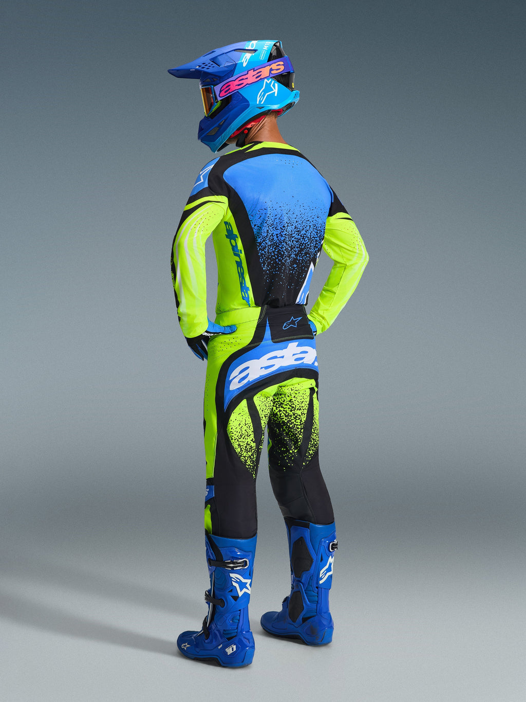Pilota di motocross visto da dietro con abbigliamento da gara completo Alpinestars, Maglia Techstar Nomur 2026 in giallo fluo e blu, pantaloni coordinati gialli e neri con dettagli blu, casco blu con logo Alpinestars rosa e stivali blu, scatto in studio che mette in risalto il design posteriore e la vestibilità atletica.