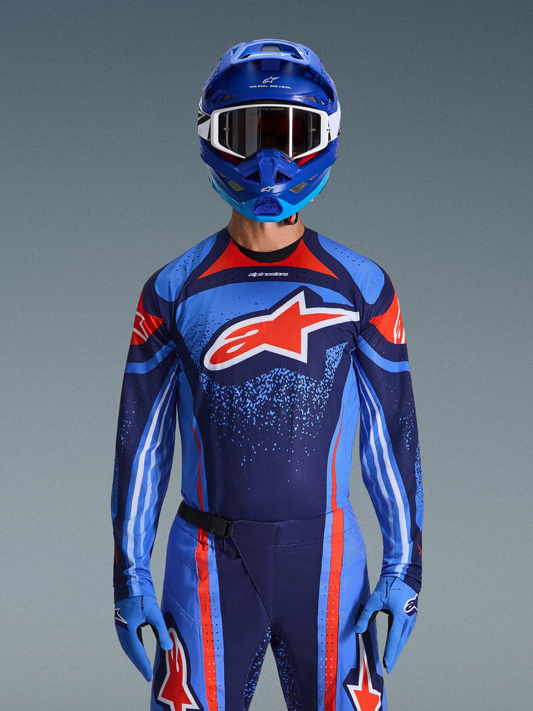 Pilota di motocross che indossa la maglia Alpinestars 2026 Techstar Nomur in blu scuro, arancione e blu UCLA, pantaloni coordinati e guanti blu, casco blu con occhiali bianchi, grande logo Alpinestars rosso e bianco sul petto, abbigliamento tecnico coordinato per corse fuoristrada.