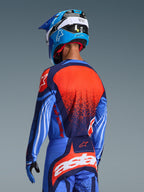 Pilota di motocross visto di spalle che indossa la maglia Alpinestars Techstar Nomur 2026 e i pantaloni coordinati, Dark Navy e UCLA Blue con vivaci accenti arancioni, con una grande grafica a stella e ventilazione tagliata al laser sulla maglia, abbinati a un casco blu e bianco per un kit da corsa off-road completo