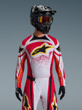 Alpinestars Techstar Nomur Jersey 2026, maglia da motocross, rosso, bianco e nero, con un grande logo A-star nero sul petto e accenti gialli, design con effetto splatter rosso, indossata con casco nero e attrezzatura off-road coordinata