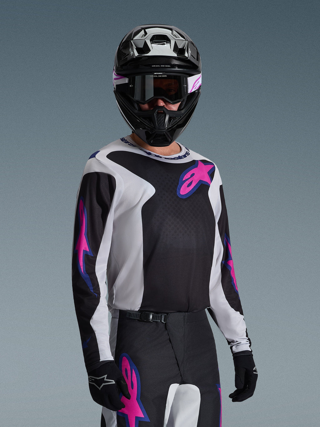 Pilota di motocross che indossa la maglia Alpinestars Fluid Grid 2026, nero, grigio chiaro e viola, con grandi loghi a stella rosa e viola, abbinata a pantaloni coordinati, casco nero e occhiali, set completo di abbigliamento da gara off-road