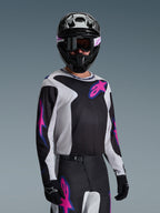 Pilota di motocross che indossa la maglia Alpinestars Fluid Grid 2026, nero, grigio chiaro e viola, con grandi loghi a stella rosa e viola, abbinata a pantaloni coordinati, casco nero e occhiali, set completo di abbigliamento da gara off-road