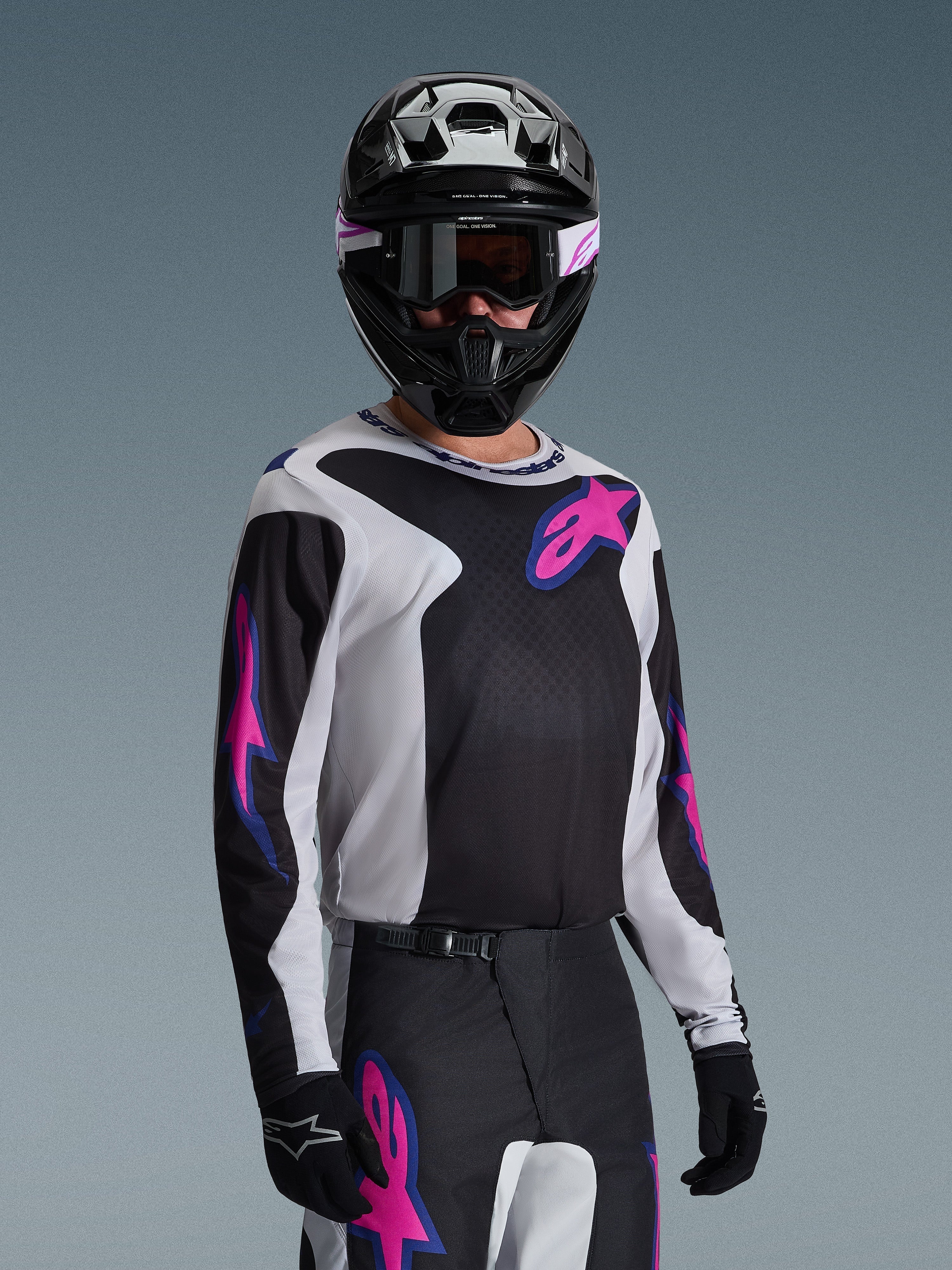 Pilota di motocross che indossa la maglia Alpinestars Fluid Grid 2026, nero, grigio chiaro e viola, con grandi loghi a stella rosa e viola, abbinata a pantaloni coordinati, casco nero e occhiali, set completo di abbigliamento da gara off-road