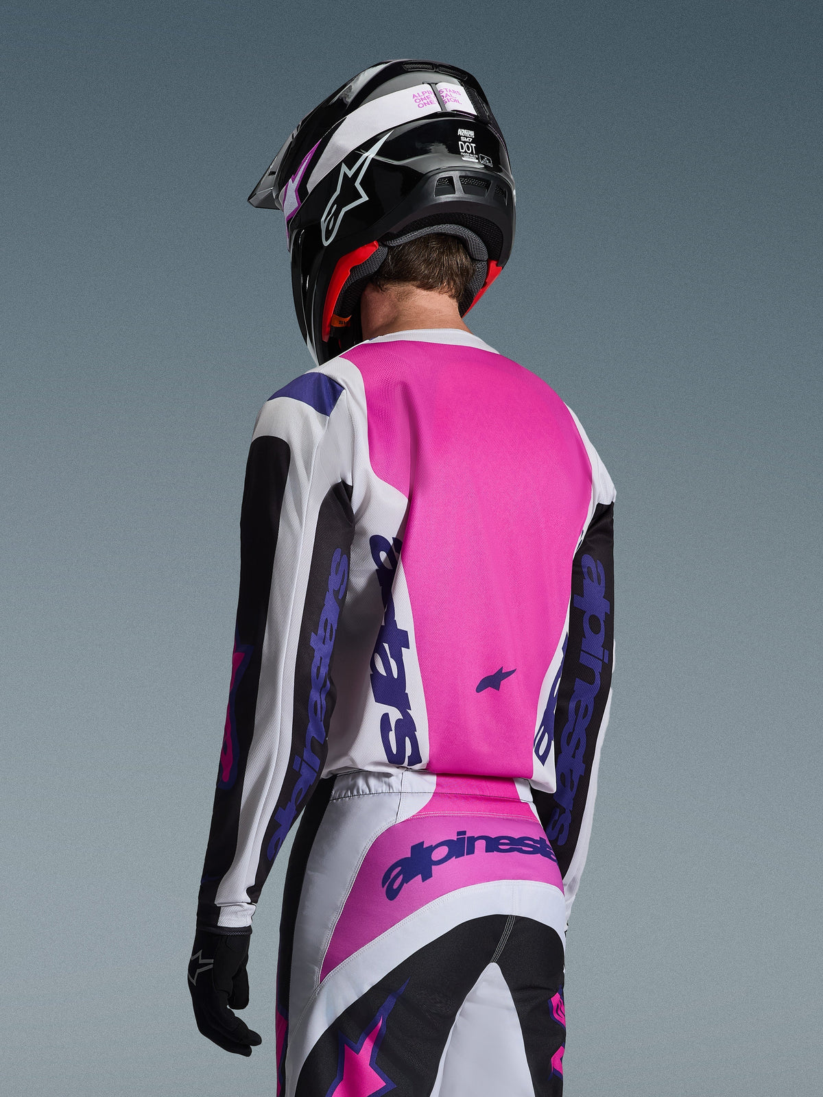 Pilota di motocross visto di spalle che indossa la maglia Alpinestars 2026 Fluid Grid in nero, bianco e viola, pantaloni coordinati con branding audace, casco nero con accenti rosa e guanti neri, abbigliamento coordinato completo per prestazioni fuoristrada.