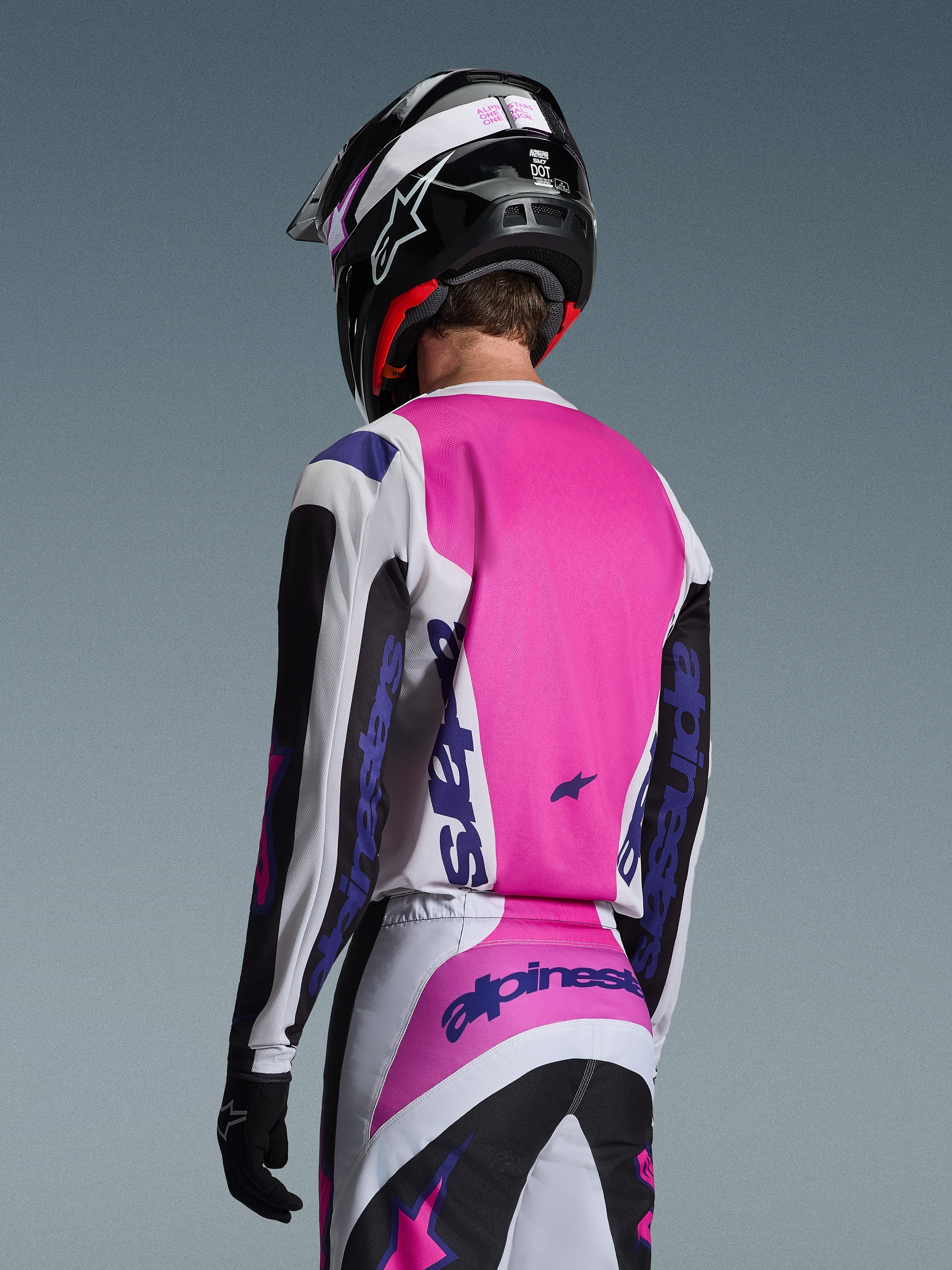 Pilota di motocross visto di spalle che indossa la maglia Alpinestars 2026 Fluid Grid in nero, bianco e viola, pantaloni coordinati con branding audace, casco nero con accenti rosa e guanti neri, abbigliamento coordinato completo per prestazioni fuoristrada.