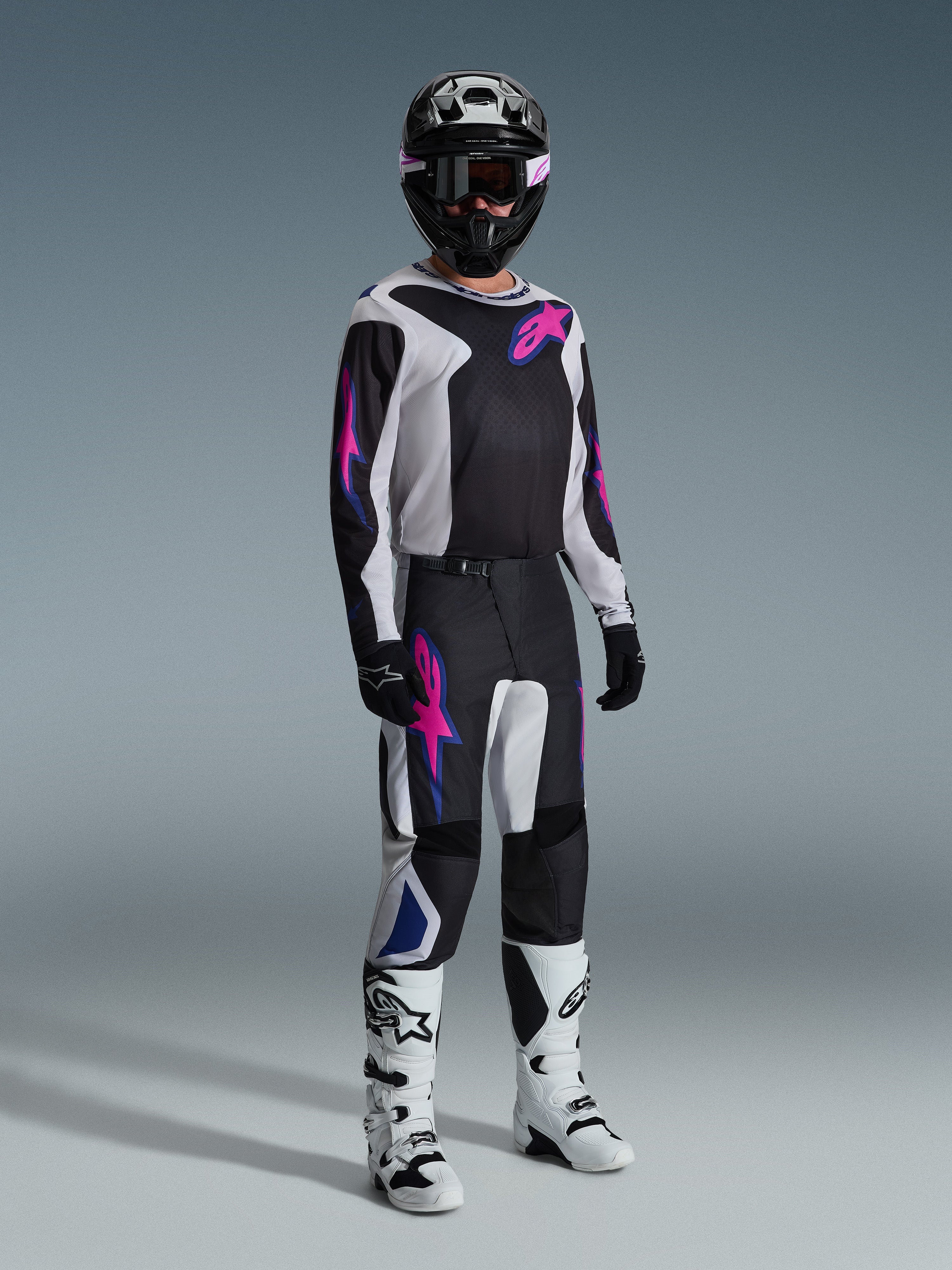 Alpinestars 2026 Fluid Grid Gear Set, pilota di motocross con maglia e pantaloni neri e bianchi con accenti rosa e blu, casco nero, guanti neri e stivali bianchi, completo da gara su sfondo grigio