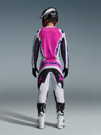 Pilota di motocross visto da dietro che indossa il set Alpinestars 2026 Fluid Grid, con maglia rosa acceso e bianca con maniche nere, pantaloni bianchi coordinati con pannelli neri e rosa, logo Alpinestars viola, stivali da gara bianchi e neri e casco abbinato, progettato per prestazioni off-road.