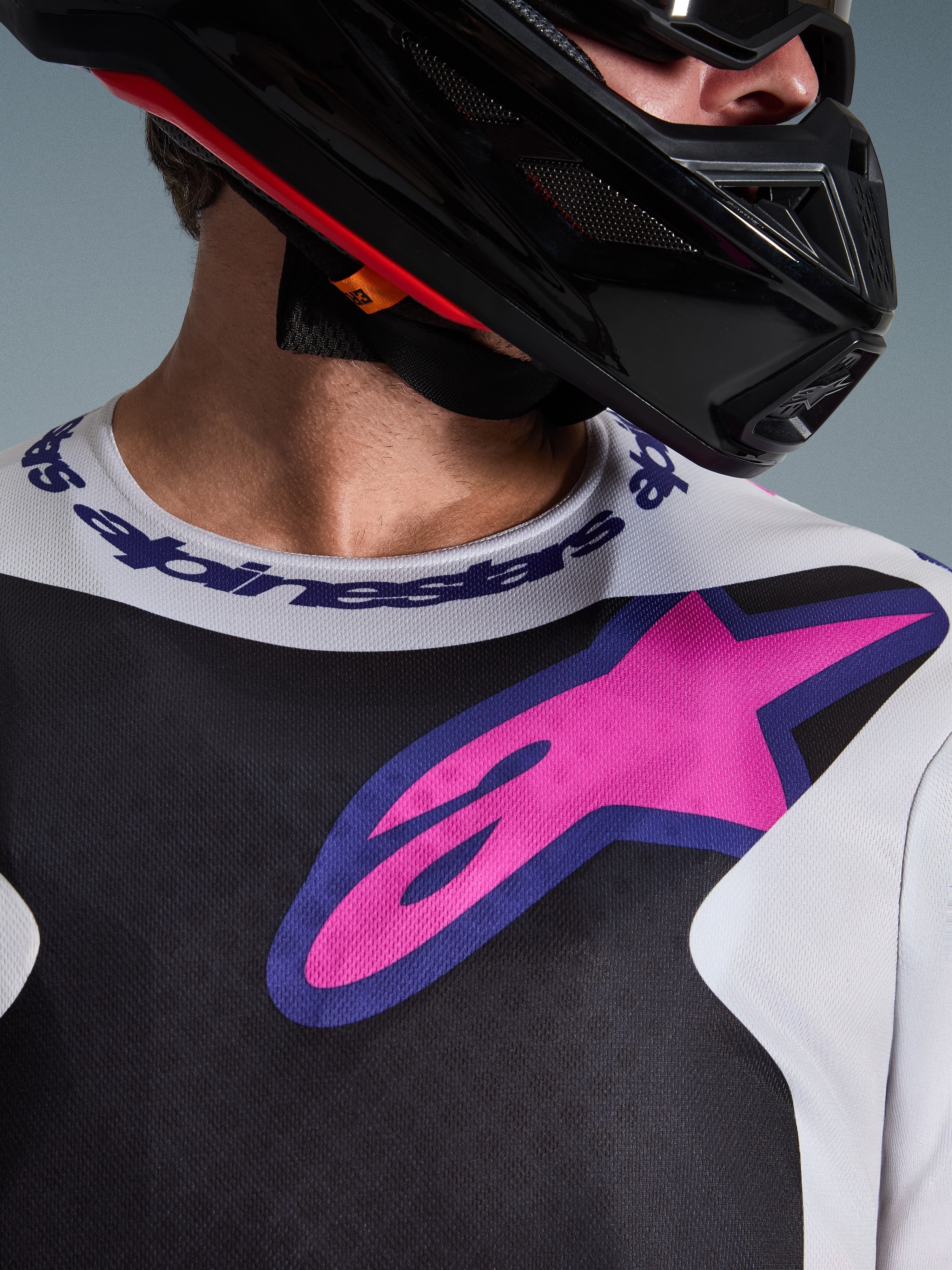 Alpinestars 2026 Fluid Grid Jersey, maglia da motocross, nero, grigio chiaro e viola, primo piano di petto e collo con grande logo a stella rosa, colletto sportivo logato, indossata con casco nero, abbigliamento tecnico da fuoristrada