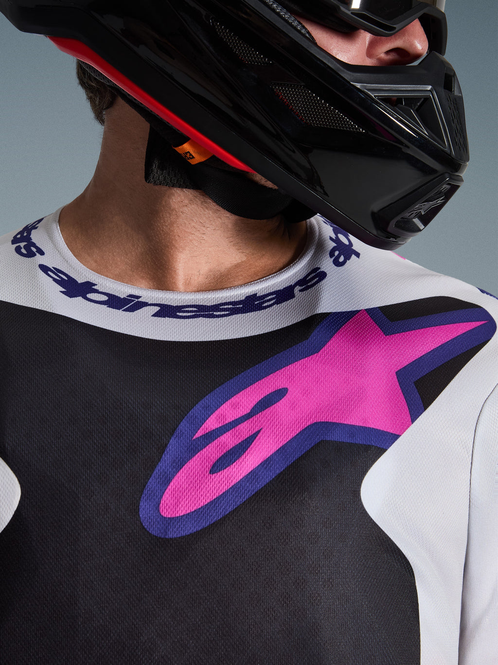 Primo piano della maglia Alpinestars Fluid Grid 2026, maglia da motocross, nero, grigio chiaro e viola, con vivace logo a stella rosa-viola sul petto e colletto sportivo logato, indossata con un casco da motocross nero