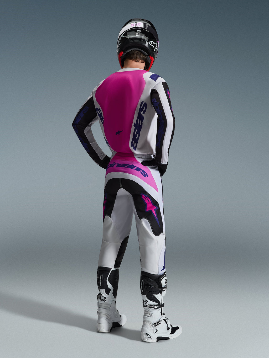 Set completo Alpinestars Fluid Grid 2026, pilota di motocross visto di spalle, maglia bianca con pannello posteriore rosa e branding viola, pantaloni coordinati bianchi e neri con accenti rosa, stivali bianchi e neri e casco nero, abbigliamento coordinato per corse fuoristrada