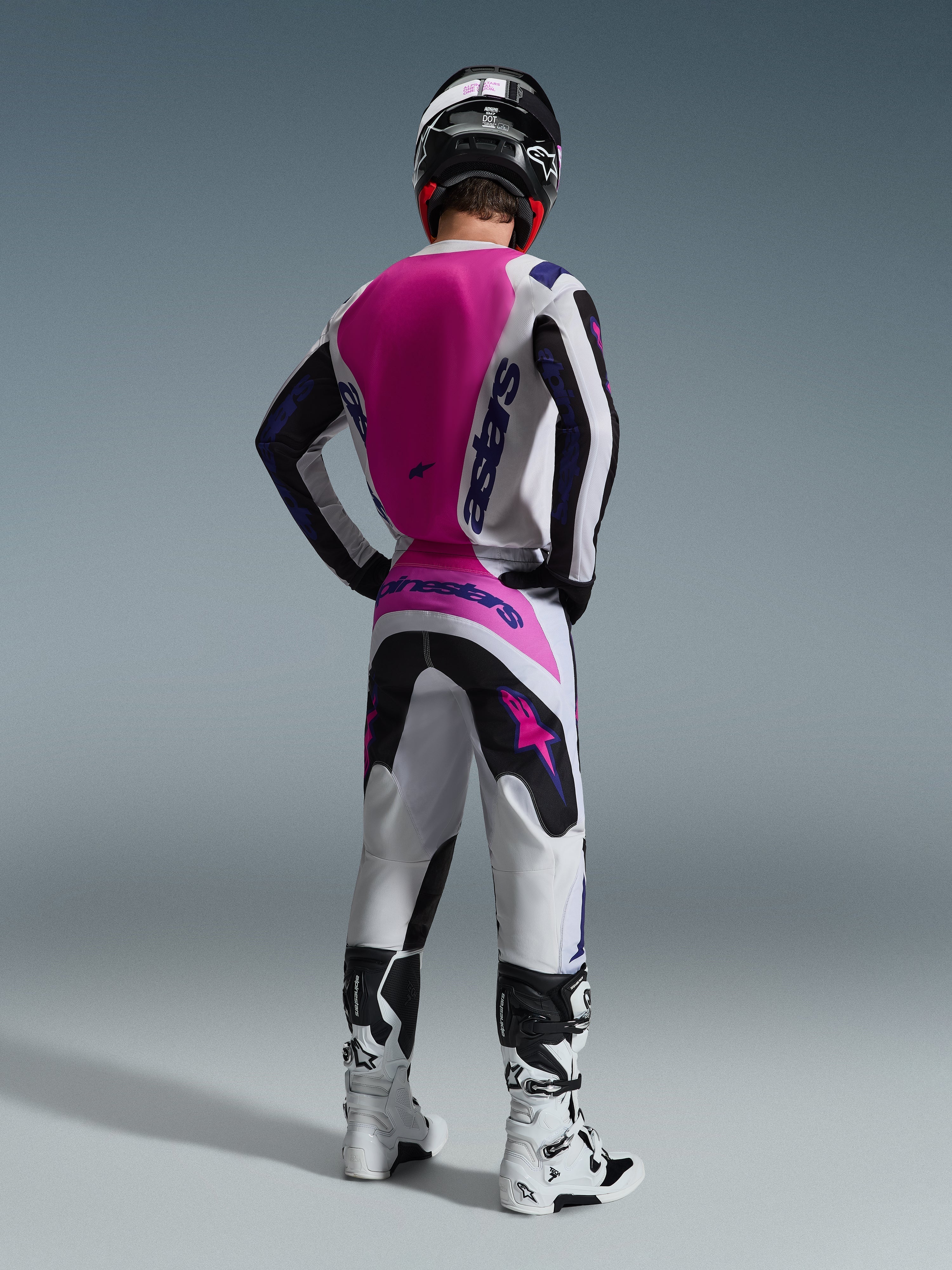 Set completo Alpinestars Fluid Grid 2026, pilota di motocross visto di spalle, maglia bianca con pannello posteriore rosa e branding viola, pantaloni coordinati bianchi e neri con accenti rosa, stivali bianchi e neri e casco nero, abbigliamento coordinato per corse fuoristrada