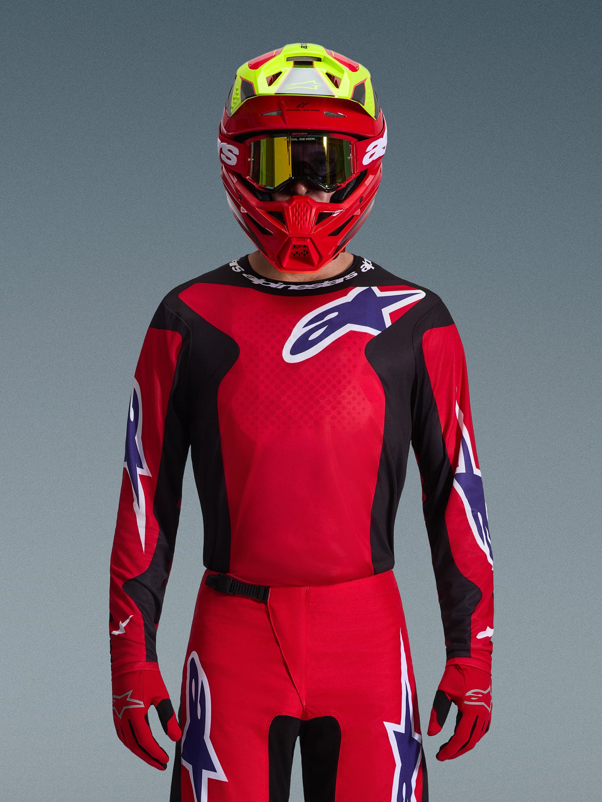 Alpinestars 2026 Fluid Grid Jersey, maglia da motocross, rosso, nero e viola, indossata da un pilota con casco rosso e giallo neon e occhiali dorati, con grandi loghi a stella sul petto e sulle maniche, abbigliamento coordinato per corse fuoristrada
