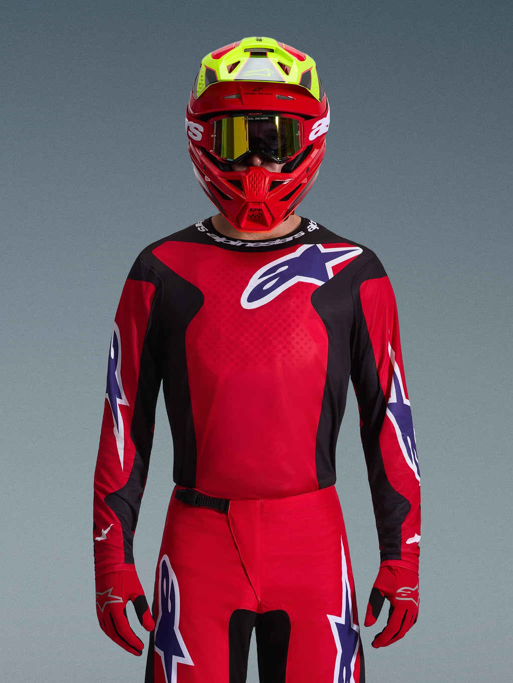 Alpinestars 2026 Fluid Grid Jersey, maglia da motocross, rosso, nero e viola, indossata da un pilota con casco rosso e giallo neon e occhiali dorati, con grandi loghi a stella sul petto e sulle maniche, abbigliamento coordinato per corse fuoristrada