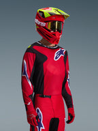 Pilota di motocross con completo Alpinestars Fluid Grid 2026, maglia rossa e nera con dettagli del logo bianchi e blu, pantaloni coordinati rossi e neri, casco rosso con tocchi gialli e maschera dorata, equipaggiamento da gara fuoristrada coordinato