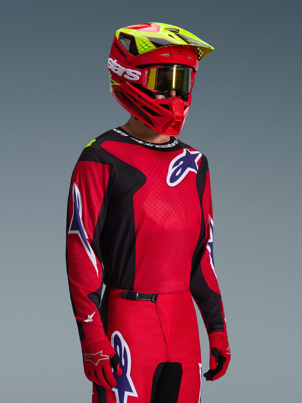 Pilota di motocross con maglia Alpinestars Fluid Grid 2026 in rosso, nero e viola, coordinata con pantaloni da motocross abbinati, casco rosso con accenti giallo neon, maschera dorata e guanti rossi, completo da gara off-road.