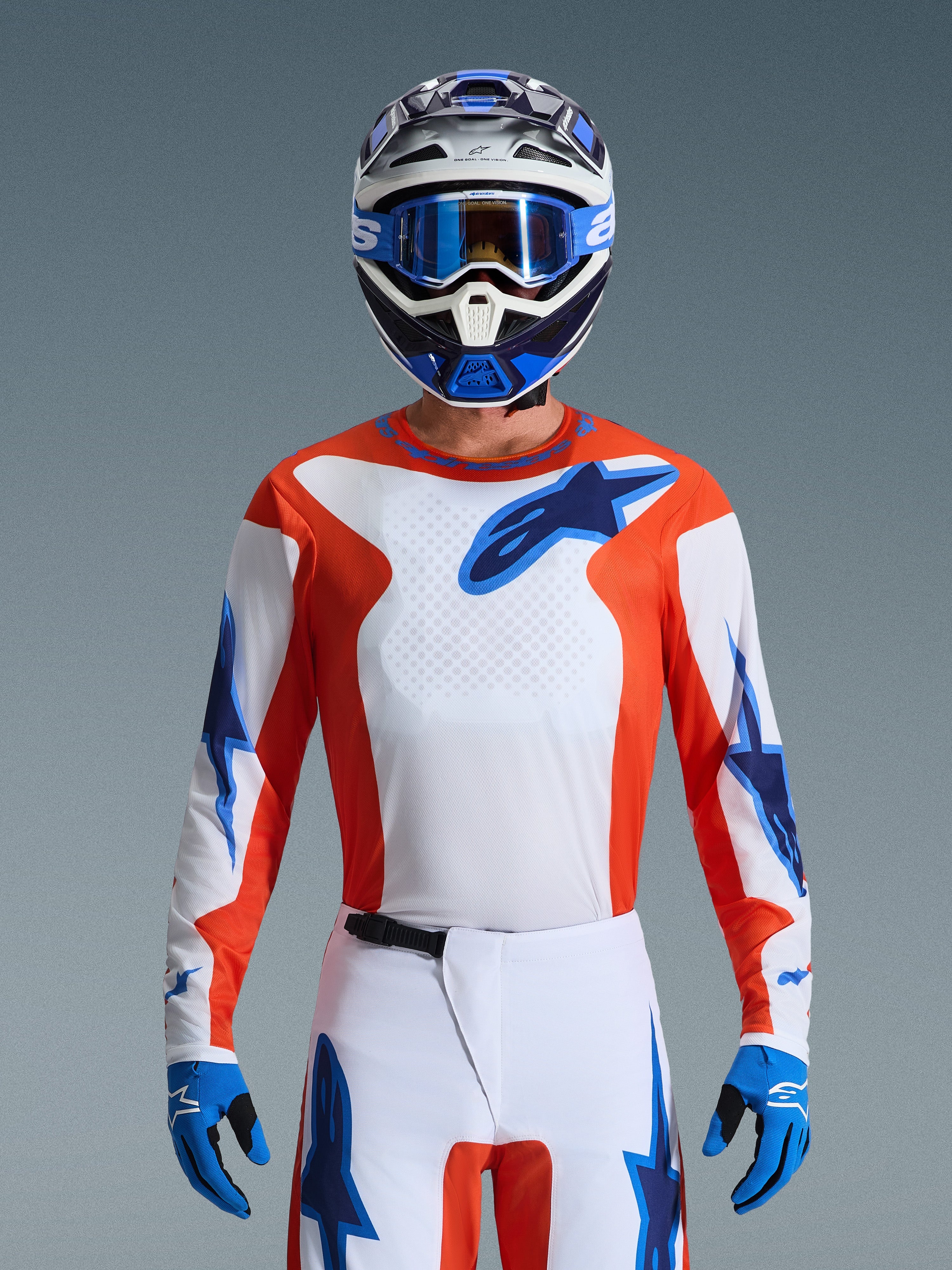 Pilota di motocross che indossa la maglia Fluid Grid 2026 in Orange Ucla Blue, caratterizzata da petto bianco con pannelli laterali arancioni e loghi Alpinestars blu, coordinata con pantaloni bianchi e blu abbinati, guanti blu e un casco bianco e blu per un look professionale da corsa off-road.