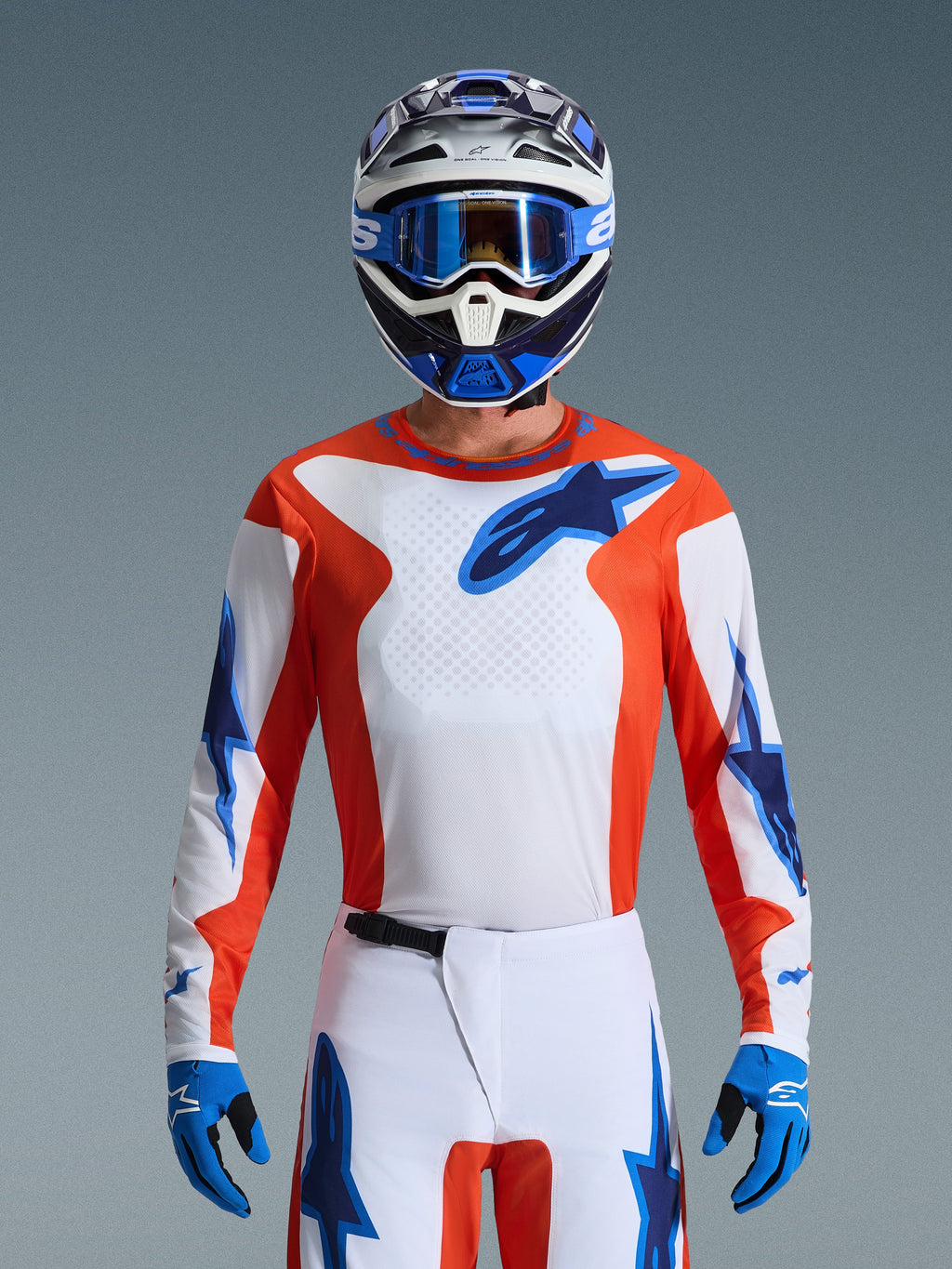 Alpinestars 2026 Fluid Grid Gear Set, pilota di motocross con maglia arancione e bianca e loghi a stella blu, pantaloni bianchi con dettagli blu, casco coordinato bianco e blu con occhiali e guanti blu, abbigliamento completo da corsa fuoristrada