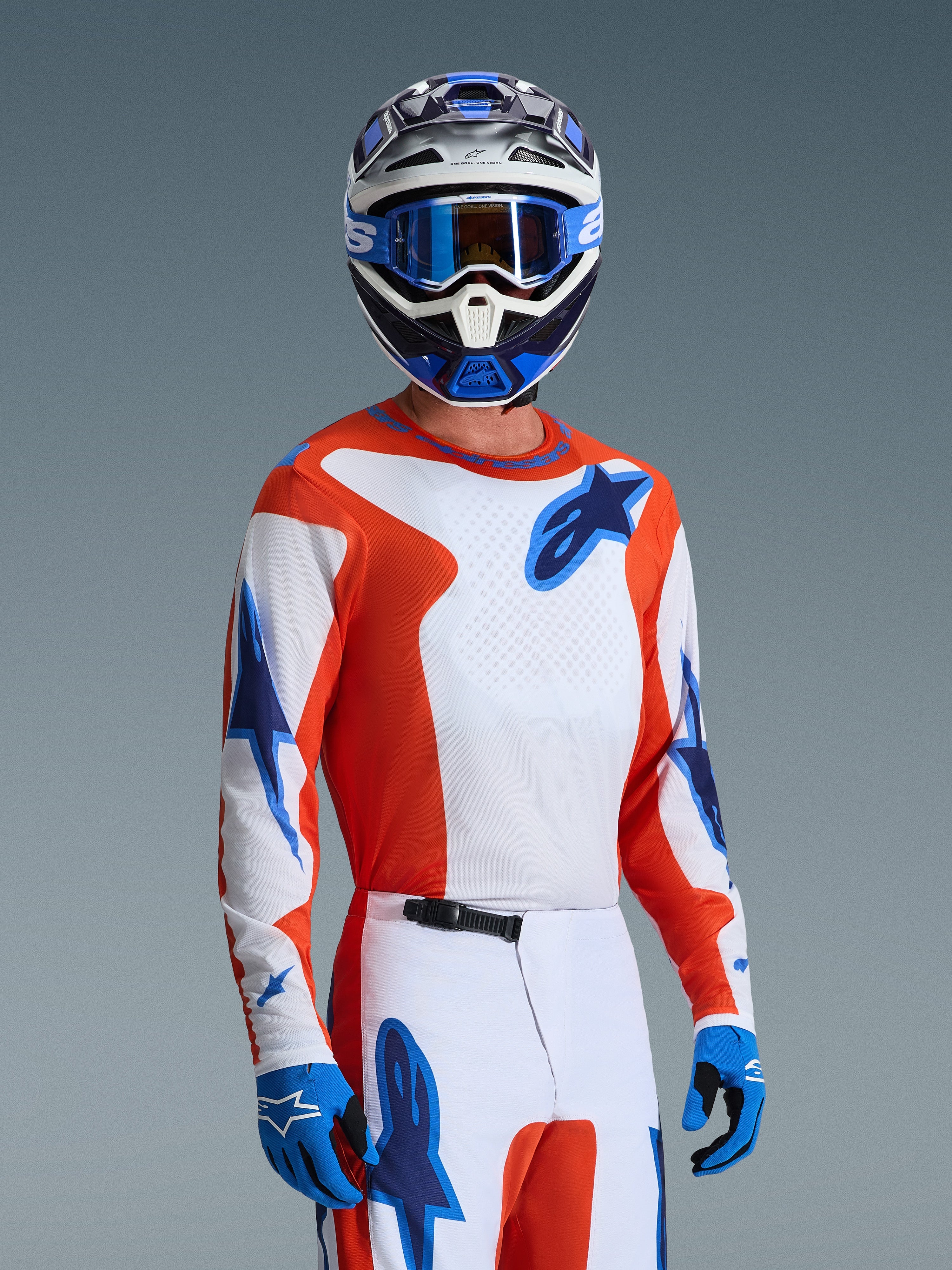 Alpinestars Maglia Fluid Grid 2026, maglia da motocross in Orange UCLA Blue, indossata da un pilota con pantaloni bianchi e arancioni coordinati, casco bianco e blu, maschera blu e guanti blu, completo da gara off-road