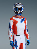 Pilota di motocross con set completo Alpinestars 2026 Fluid Grid, maglia arancione e bianca con loghi a stella blu, pantaloni bianchi e arancioni con dettagli blu, guanti blu, casco bianco e blu con occhiali blu, abbigliamento tecnico coordinato per prestazioni off-road