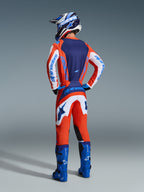 Pilota di motocross visto di spalle che indossa maglia e pantaloni Fluid Grid 2026 in arancione e blu UCLA, con maniche bianche e marchio Alpinestars blu, stivali da motocross blu acceso e casco coordinato, equipaggiamento completo per corse fuoristrada.