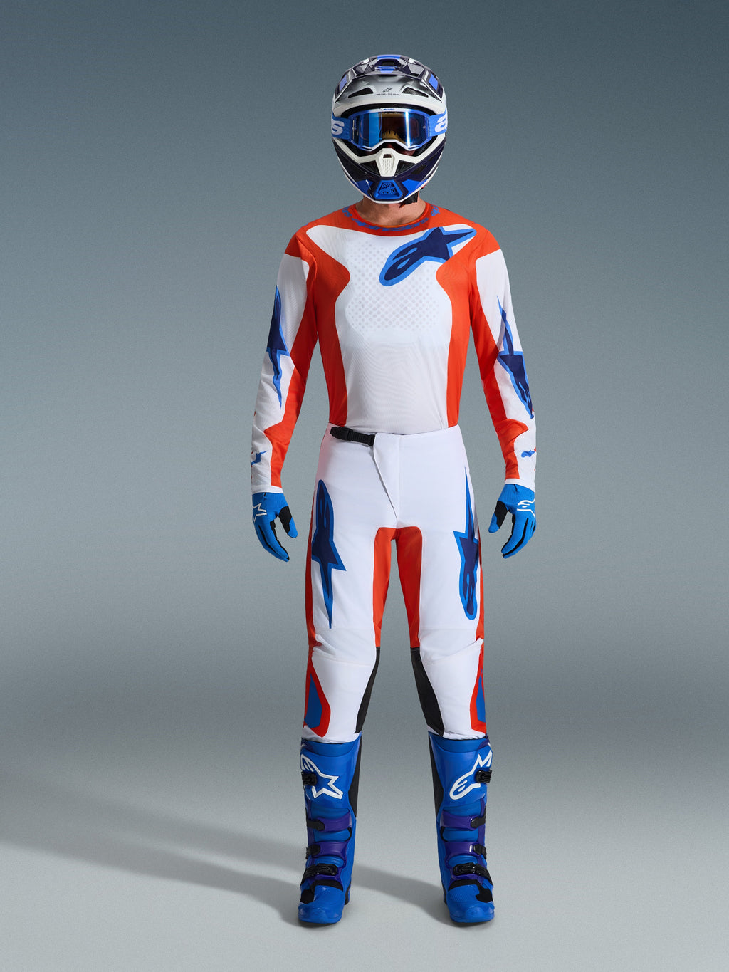Set Completo Alpinestars 2026 Fluid Grid, pilota di motocross con abbigliamento completo, maglia e pantaloni bianchi con dettagli arancioni e blu UCLA, casco coordinato bianco e blu con maschera blu, guanti blu e stivali da motocross blu, completo da corsa off-road coordinato