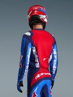 Vista posteriore di un pilota di motocross che indossa la maglia Alpinestars 2026 Fluid Grid in Blu e Rosso, caratterizzata da loghi bianchi su maniche e schiena, abbinata a pantaloni da motocross coordinati e un casco rosso per un look da gara off-road completo