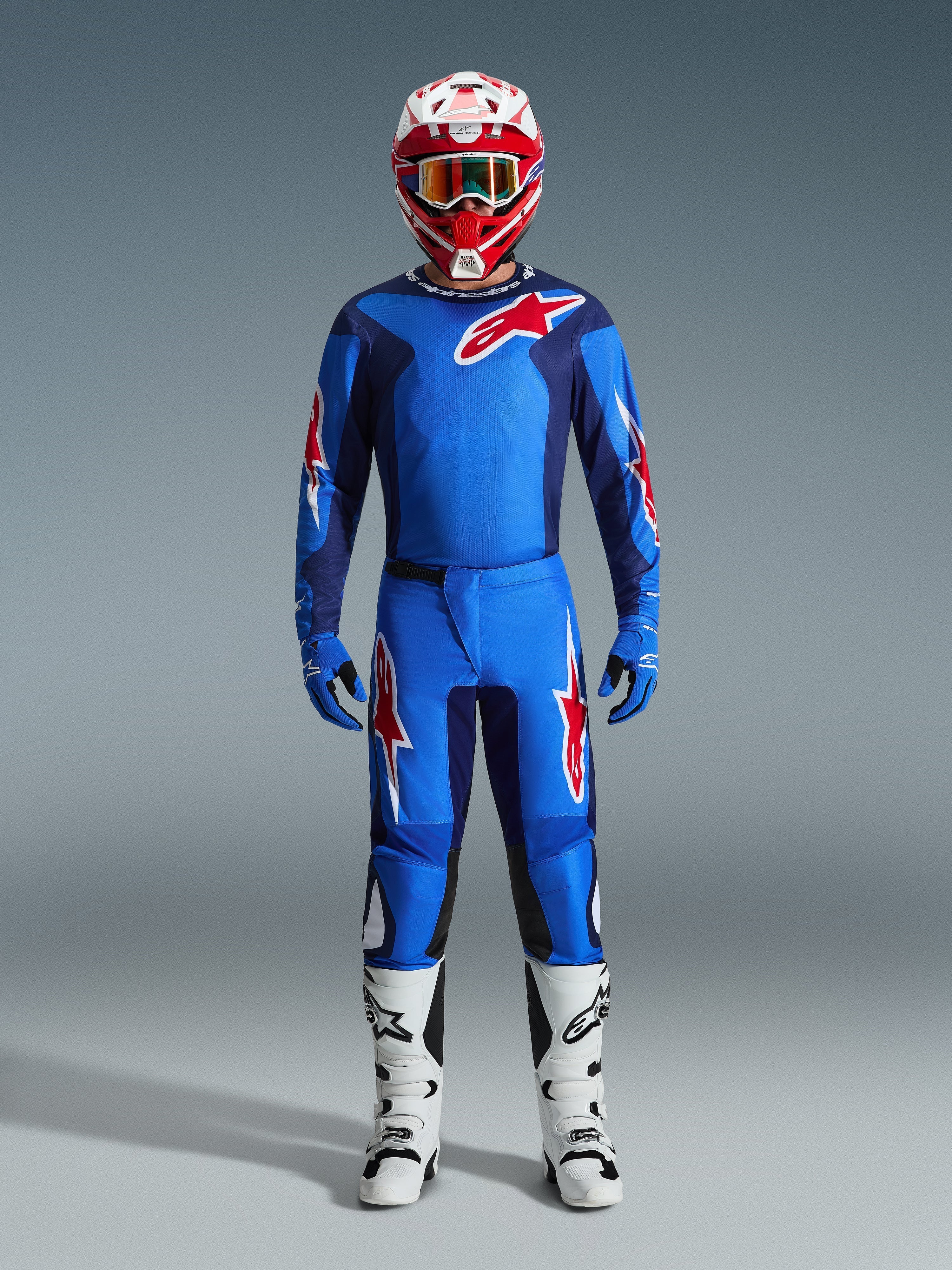 Pilota di motocross con maglia Alpinestars 2026 Fluid Grid in blu e rosso, pantaloni blu coordinati con loghi a stella bianchi e rossi, casco rosso e bianco con maschera a specchio, guanti blu e stivali da corsa bianchi e neri, set completo di abbigliamento fuoristrada.