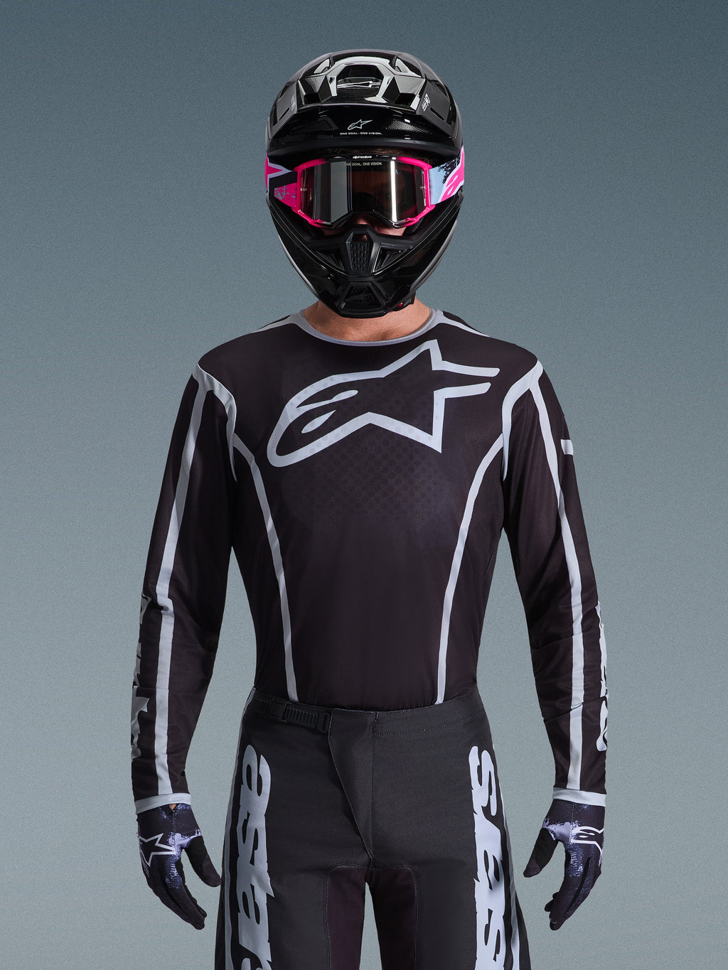 Alpinestars 2026 Fluid Apex Gear Set, maglia e pantaloni da motocross in bianco e nero, caratterizzati da un grande logo sul petto e design a linee geometriche, indossato con casco nero e occhiali rosa, abbigliamento tecnico per gare di motocross fuoristrada.