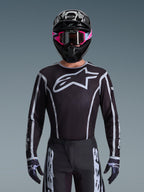 Alpinestars 2026 Fluid Apex Gear Set, maglia e pantaloni da motocross in bianco e nero, caratterizzati da un grande logo sul petto e design a linee geometriche, indossato con casco nero e occhiali rosa, abbigliamento tecnico per gare di motocross fuoristrada.
