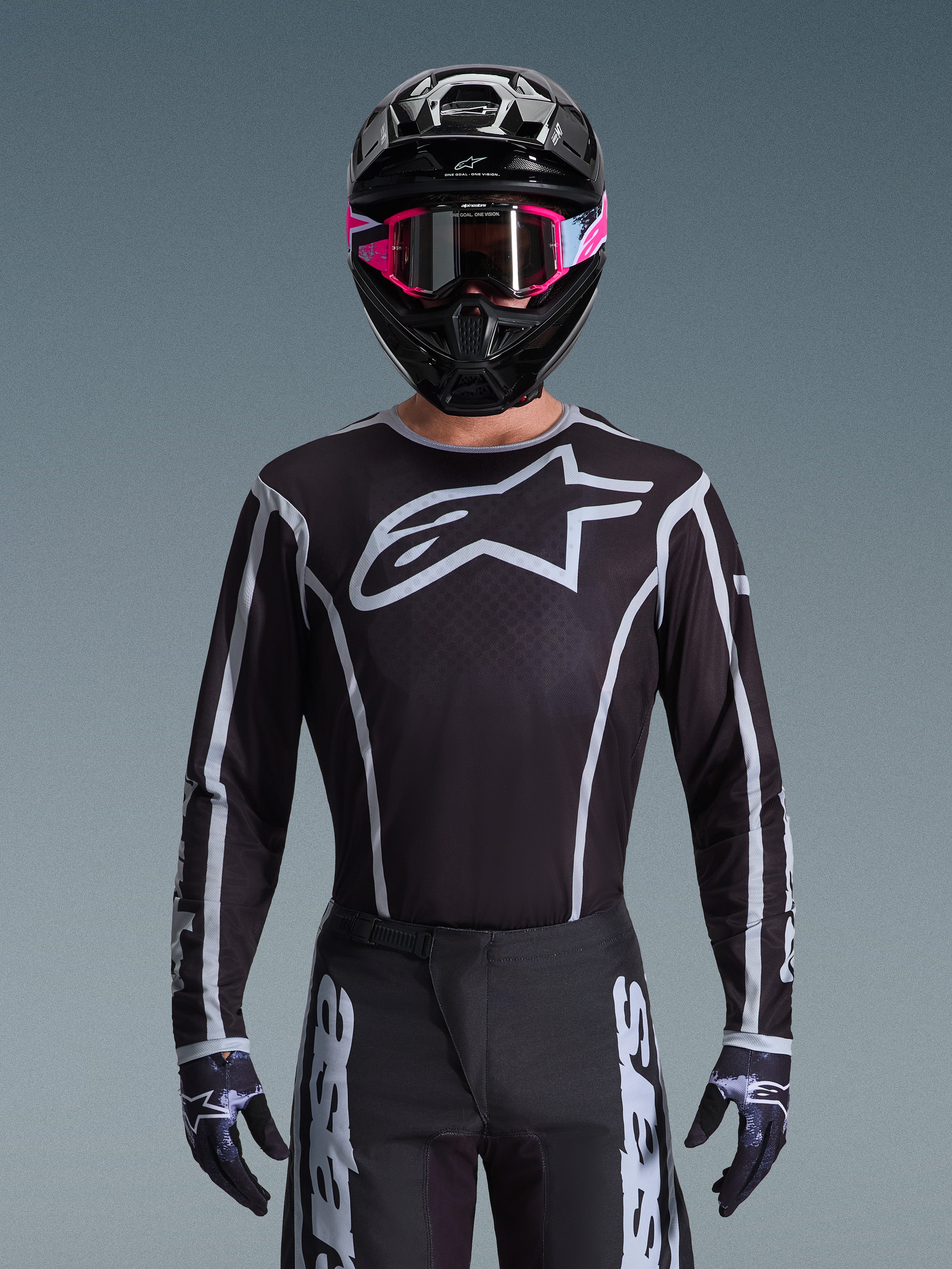 Alpinestars 2026 Fluid Apex Gear Set, maglia e pantaloni da motocross in bianco e nero, caratterizzati da un grande logo sul petto e design a linee geometriche, indossato con casco nero e occhiali rosa, abbigliamento tecnico per gare di motocross fuoristrada.