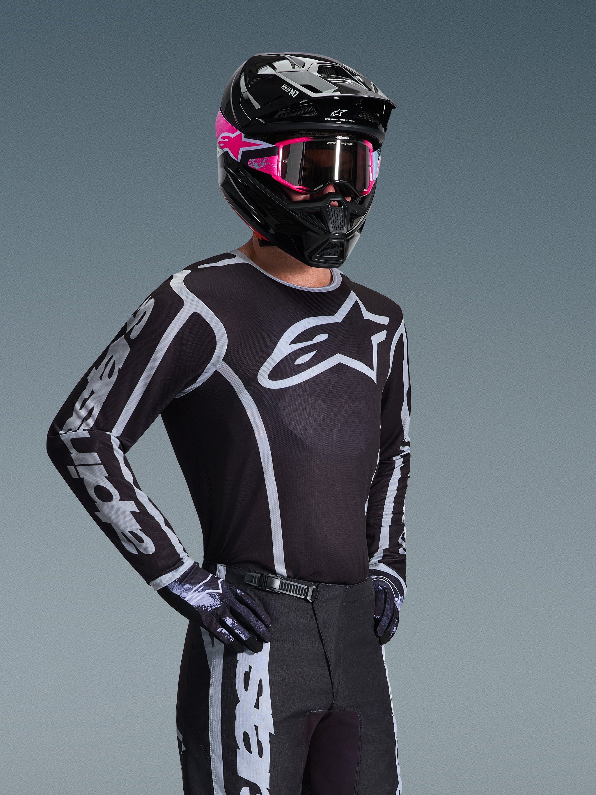 Pilota di motocross con maglia Alpinestars 2026 Fluid Apex, nero e grigio con grande logo bianco sul petto e grafiche geometriche sulle maniche, abbinata a pantaloni neri, guanti neri e casco nero con accenti rosa e maschera rosa.