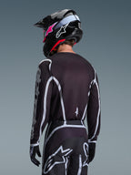 Pilota di motocross visto di spalle con maglia Alpinestars 2026 Fluid Apex in nero e grigio, pantaloni da motocross coordinati con accenti bianchi, casco nero con logo rosa, completo per corse fuoristrada