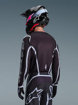 Pilota di motocross visto di spalle con maglia Alpinestars 2026 Fluid Apex in nero e grigio, pantaloni da motocross coordinati con accenti bianchi, casco nero con logo rosa, completo per corse fuoristrada