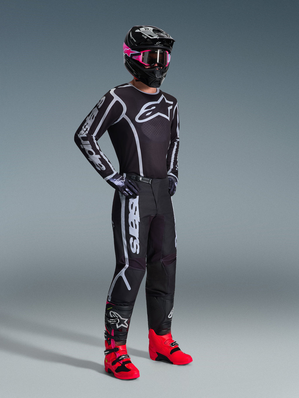 Pilota di motocross con maglia Alpinestars Fluid Apex 2026, nero e grigio, pantaloni coordinati, casco nero con maschera rosa e stivali rosso acceso, marchio bianco su petto e maniche, completo professionale da fuoristrada