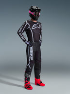 Pilota di motocross con maglia Alpinestars Fluid Apex 2026, nero e grigio, pantaloni coordinati, casco nero con maschera rosa e stivali rosso acceso, marchio bianco su petto e maniche, completo professionale da fuoristrada