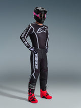 Pilota di motocross con maglia Alpinestars Fluid Apex 2026, nero e grigio, pantaloni coordinati, casco nero con maschera rosa e stivali rosso acceso, marchio bianco su petto e maniche, completo professionale da fuoristrada
