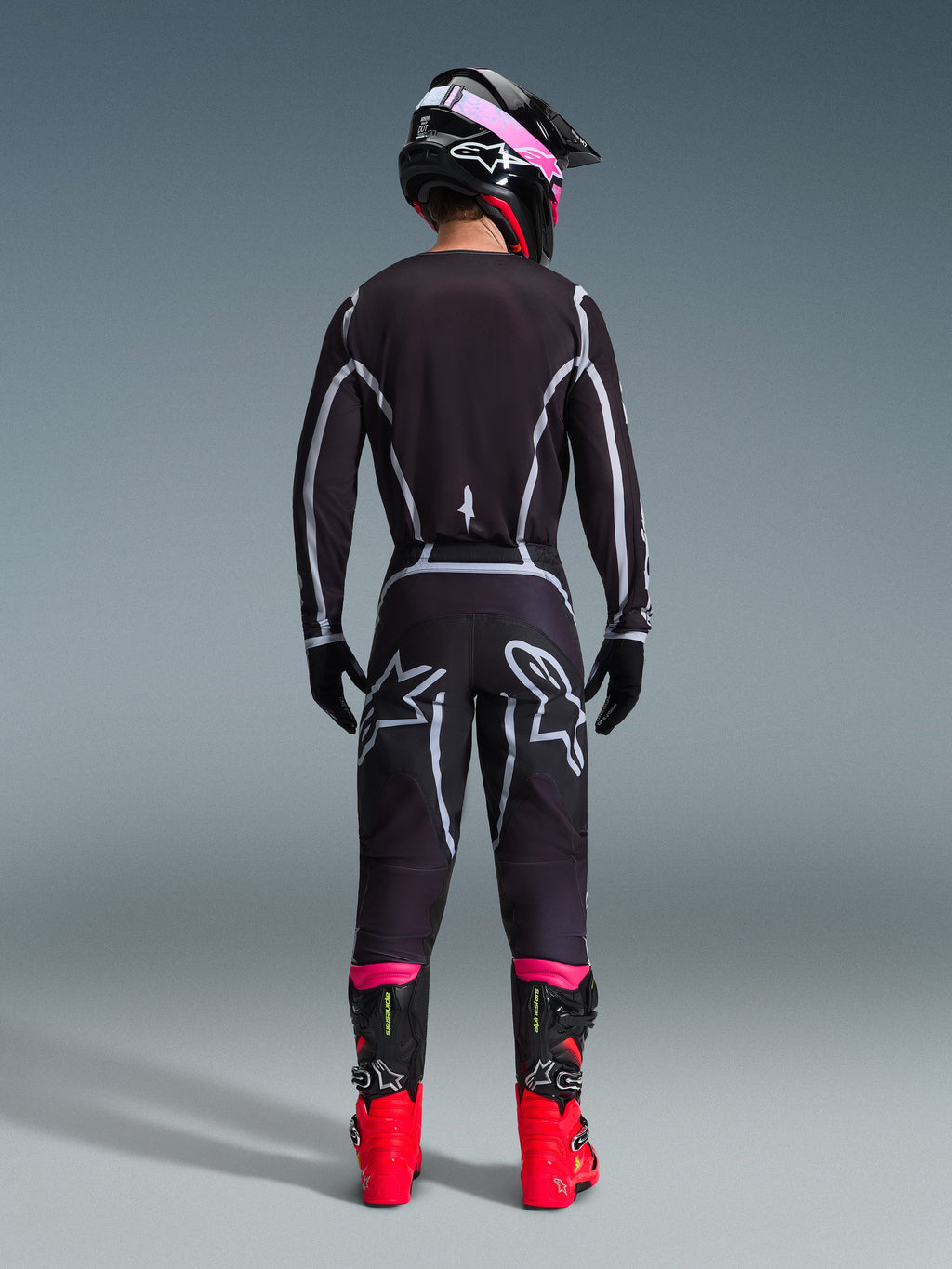 Pilota di motocross visto da dietro che indossa il completo Alpinestars 2026 Fluid Apex, maglia e pantaloni neri con linee geometriche grigie e loghi a stella, casco nero con dettagli rosa, stivali rosso acceso e neri, abbigliamento da gara motocross coordinato