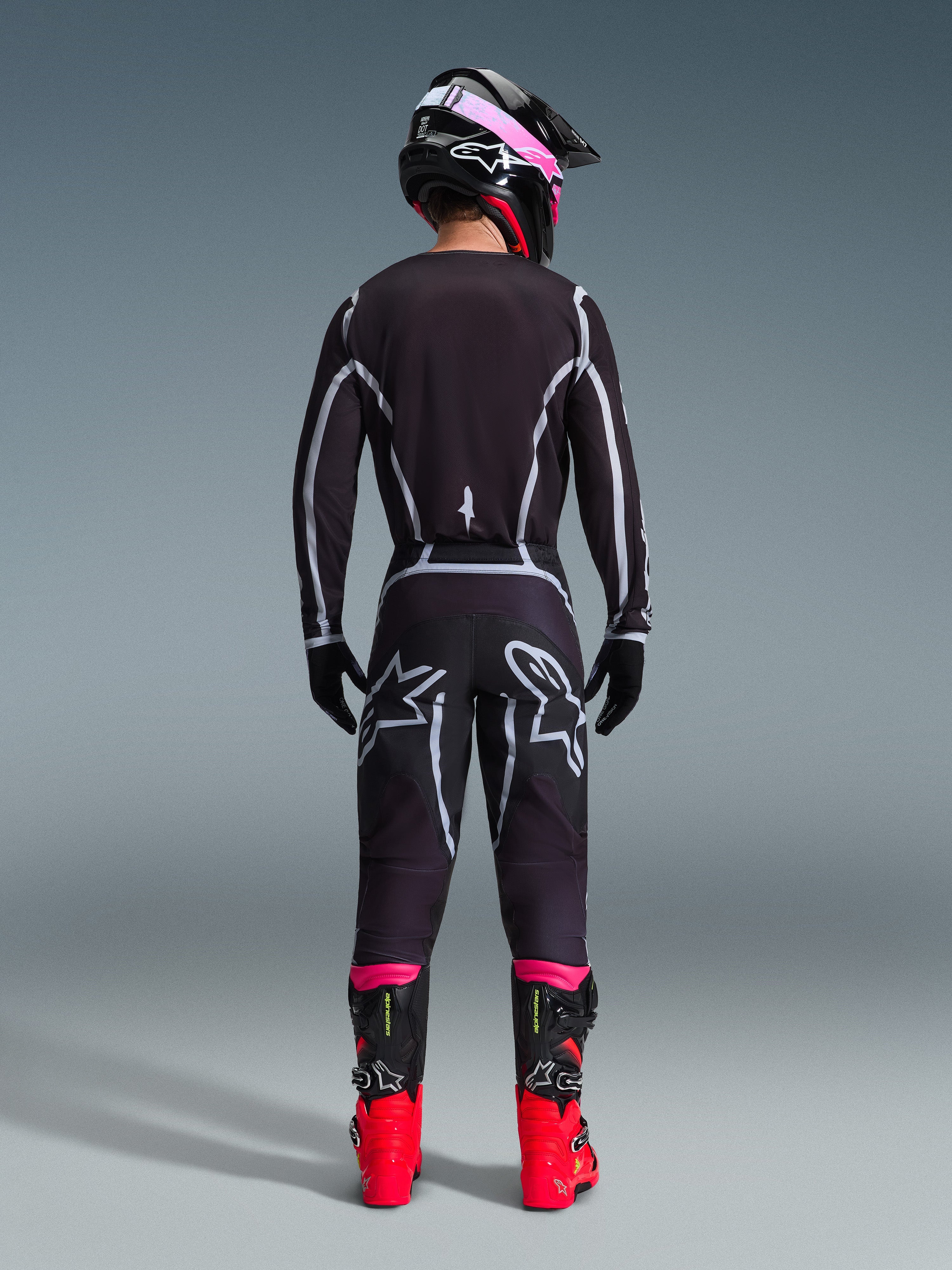 Pilota di motocross visto da dietro che indossa il completo Alpinestars 2026 Fluid Apex, maglia e pantaloni neri con linee geometriche grigie e loghi a stella, casco nero con dettagli rosa, stivali rosso acceso e neri, abbigliamento da gara motocross coordinato