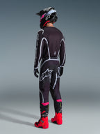 Pilota di motocross con completo Alpinestars 2026 Fluid Apex, maglia e pantaloni neri con linee geometriche bianche e grigie, vista posteriore, casco nero con dettagli rosa e stivali da corsa rosso acceso e rosa, scatto in studio su sfondo grigio.