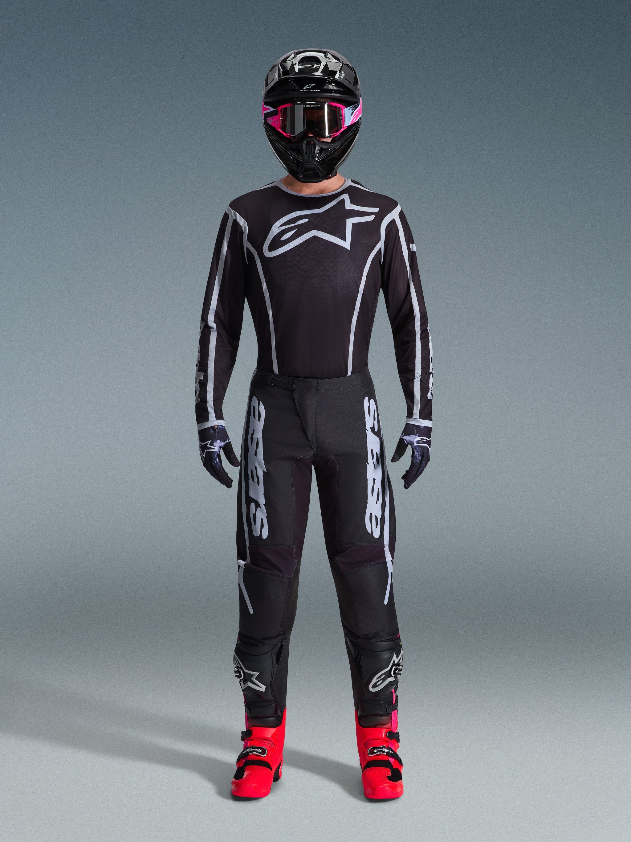 Pilota di motocross che indossa la maglia Alpinestars Fluid Apex 2026, nero e grigio, grande logo Astars bianco sul petto, linee geometriche bianche, pantaloni neri coordinati, casco nero con occhiali rosa e stivali rosa acceso, abbigliamento completo da motocross