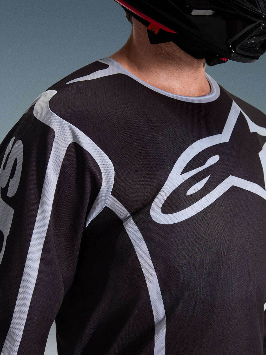 Alpinestars 2026 Fluid Apex Jersey, maglia da motocross, nero e grigio, con grande logo Alpinestars sul petto, colletto sportivo, indossata con un casco nero, abbigliamento tecnico per la guida off-road