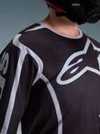 Alpinestars 2026 Fluid Apex Jersey, maglia da motocross, nero e grigio, con grande logo Alpinestars sul petto, colletto sportivo, indossata con un casco nero, abbigliamento tecnico per la guida off-road