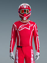 Maglia Alpinestars 2026 Fluid Apex, maglia da motocross, rossa e bianca, indossata da un pilota con casco coordinato rosso e bianco e occhiali arancioni, caratterizzata da un prominente logo bianco sul petto, vestibilità atletica, parte di un set coordinato per corse off-road